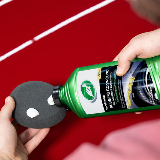 Turtle Wax rubbing compound being applied to a pad for auto body repair on a red car surface.