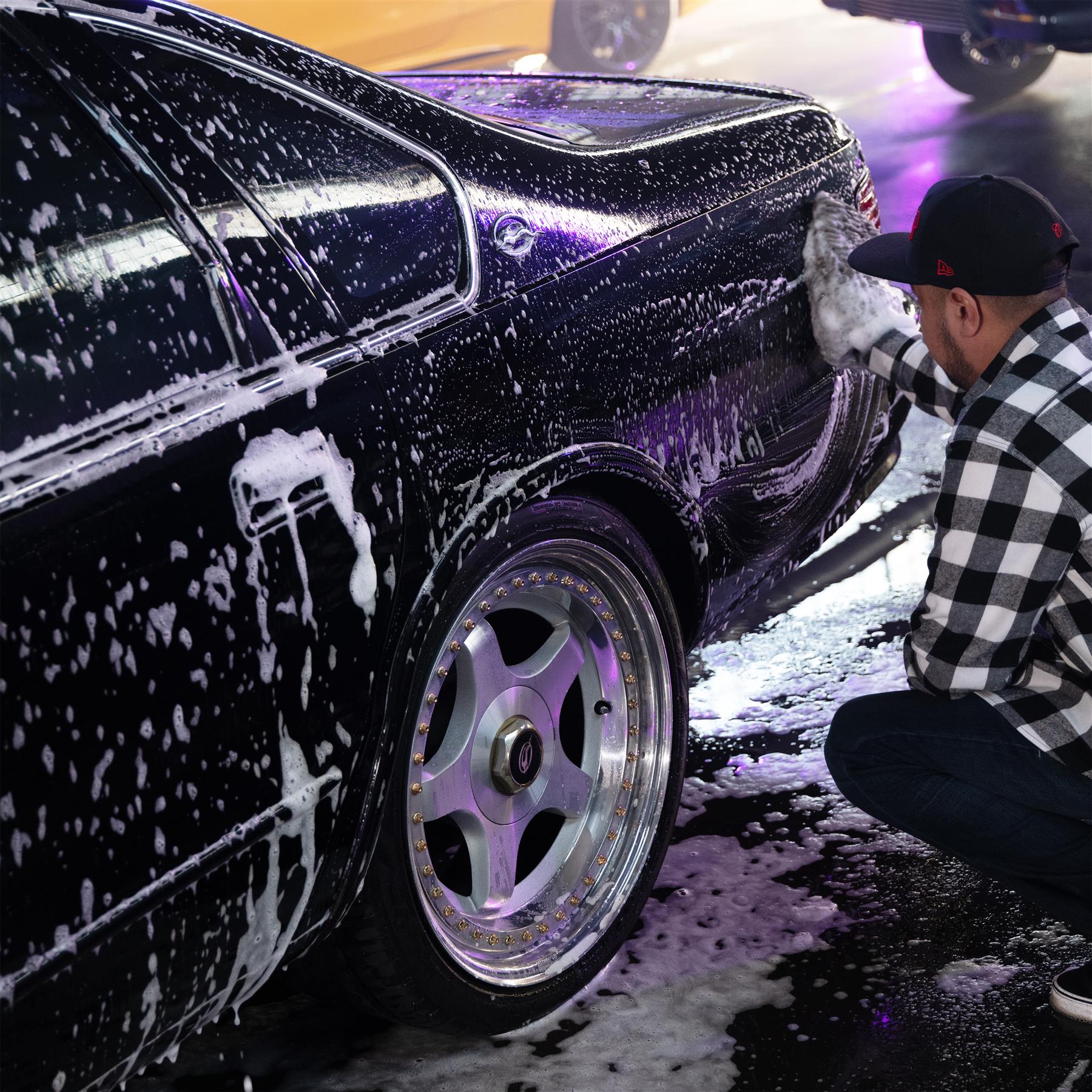 Detailer washing a dark sedan with soap suds, wearing a flannel shirt & baseball cap. Close-up on custom wheels. Car cleaning.
