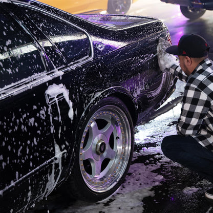 Detailer washing a dark sedan with soap suds, wearing a flannel shirt & baseball cap. Close-up on custom wheels. Car cleaning.