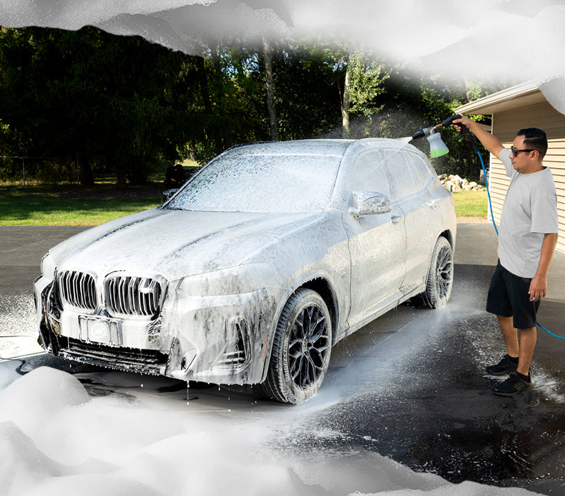 A man in a white t-shirt and black shorts washes a silver BMW SUV covered in white soap foam in a driveway, with water spraying and trees in the background.