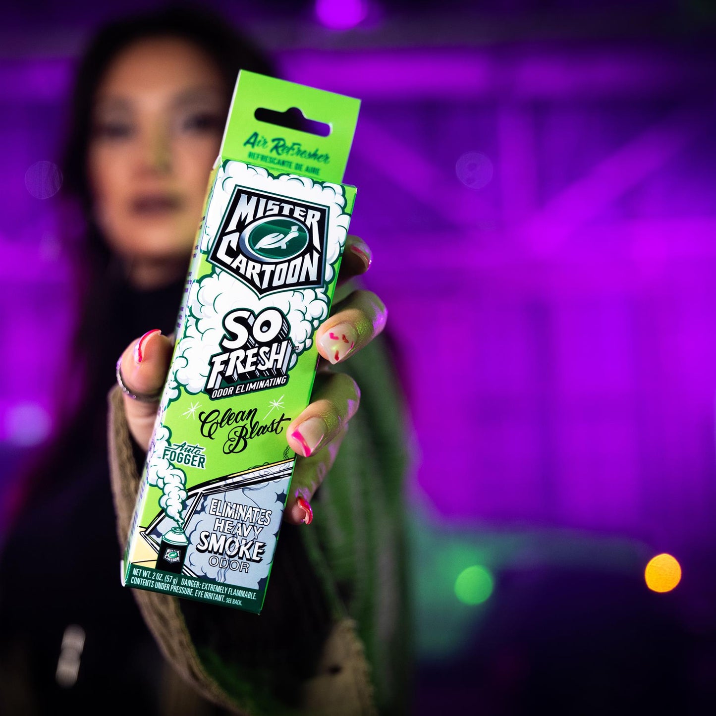 A hand displays Mister Cartoon So Fresh odor-eliminating Clean Blast Auto Fogger that eliminates heavy smoke odors in a purple scene.