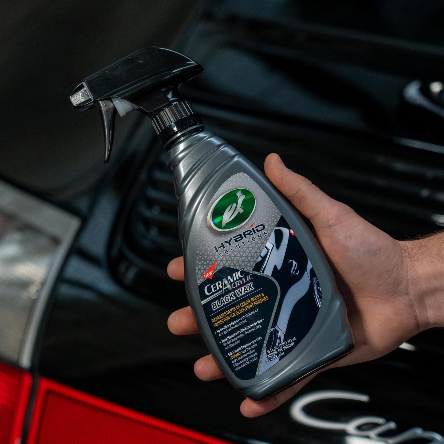 Hand holds Turtle Wax Hybrid Solutions Ceramic Acrylic Black Wax spray bottle for car detailing, with the black vehicle in background.