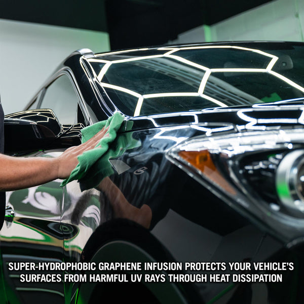 Hands using a foam applicator to spread car wax on a smooth, black surface with clear reflections.
