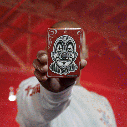 Here's a description:

Hand holding a red air freshener with a "Smile Now, Cry Later" clown graphic. The background is a blurred red.