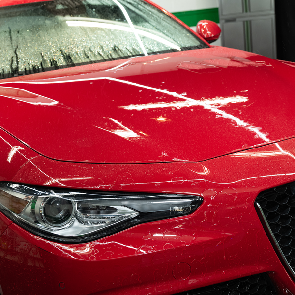 Close-up of hand applying Zip Wax Car Wash with yellow sponge, focusing on soapy texture on red vehicle's hood.