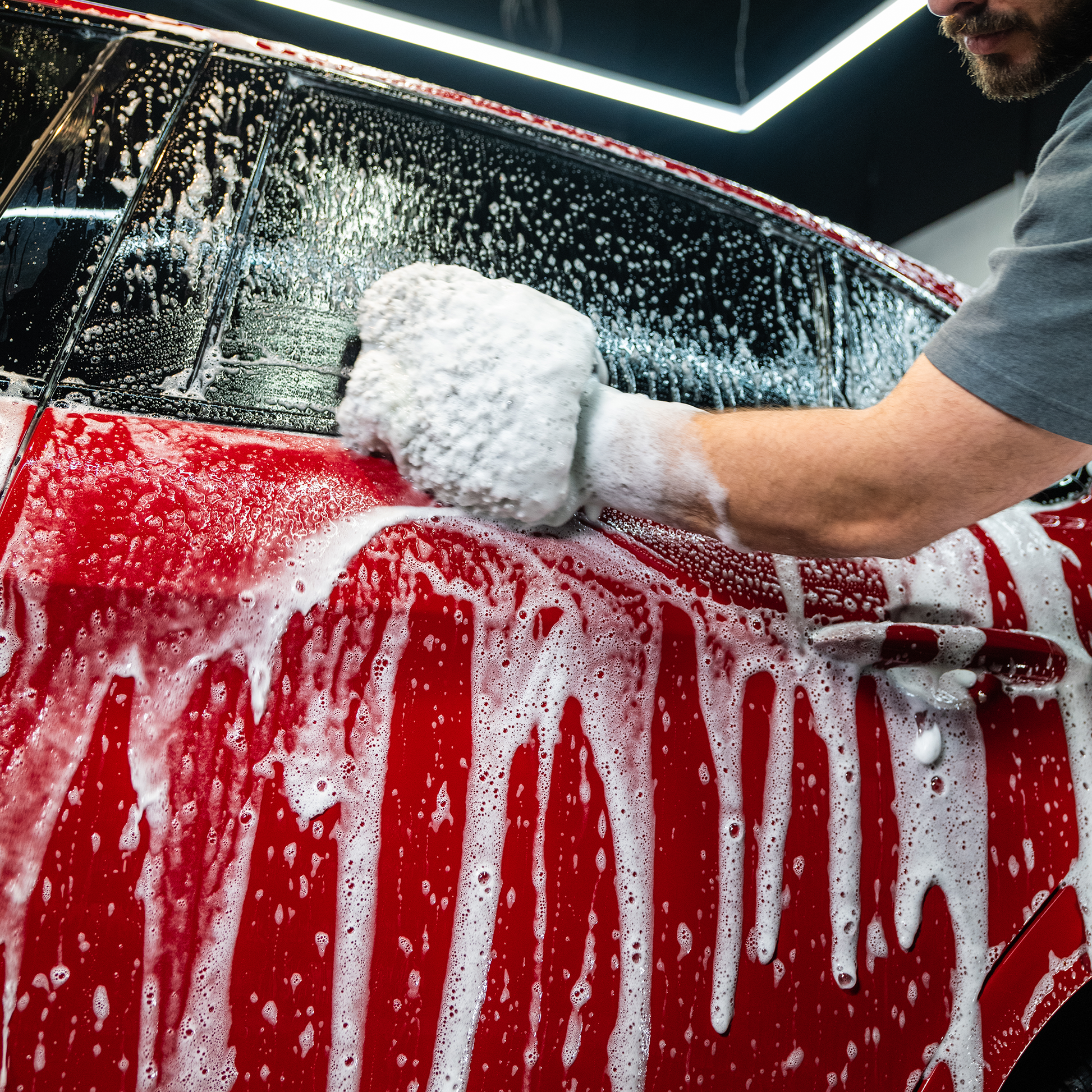 Car detailing: Red auto is being washed with soap and mitt by a man, under bright lights. Car care.