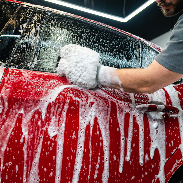 Person washing a black car with Zip Wax Car Wash, focusing on hood covered in thick suds, showing hand scrubbing with sponge.