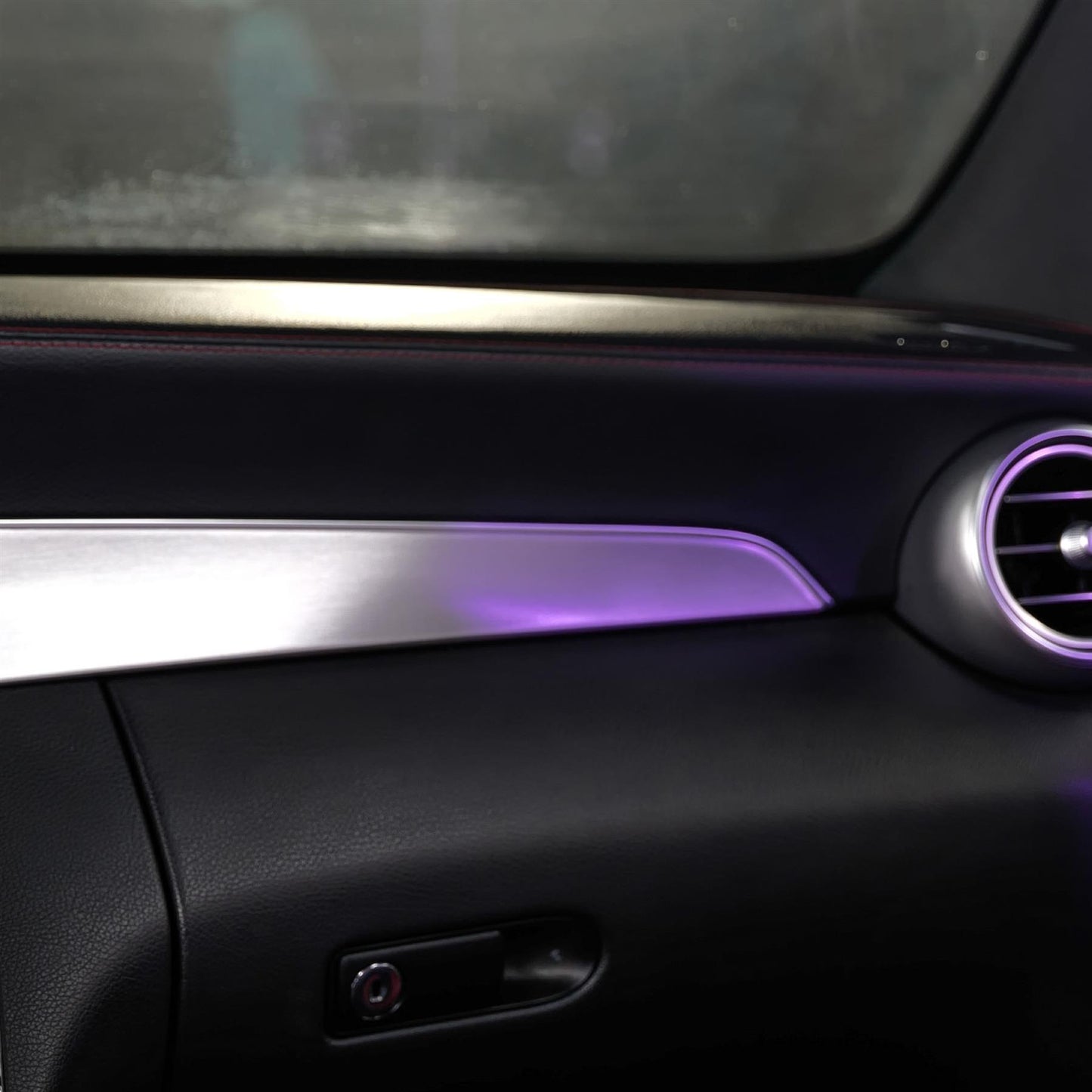 Car interior detail of a black dashboard with a silver trim strip and a round air vent. Purple lighting accentuates the elements.