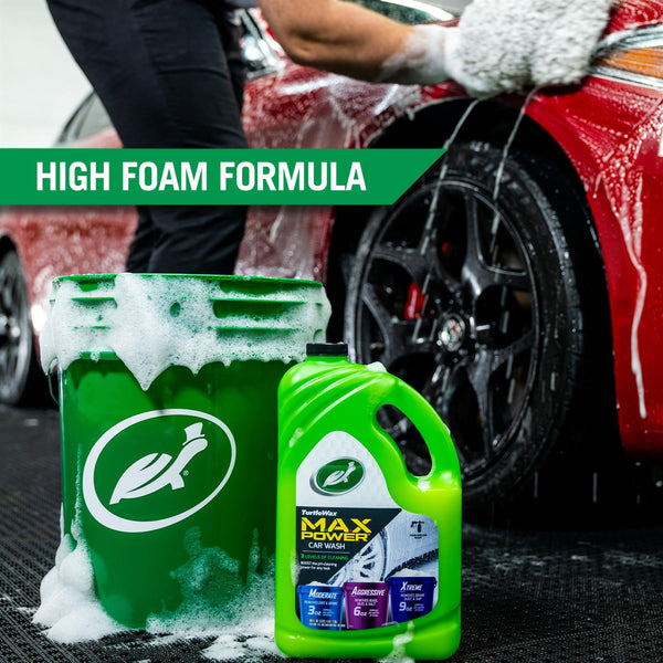 Turtle Wax MAX Power is displayed with a soapy and green 5 gallon bucket. In the background a gloved hand scrubs the surface of a red car, highlighting the high foam formula of this product.