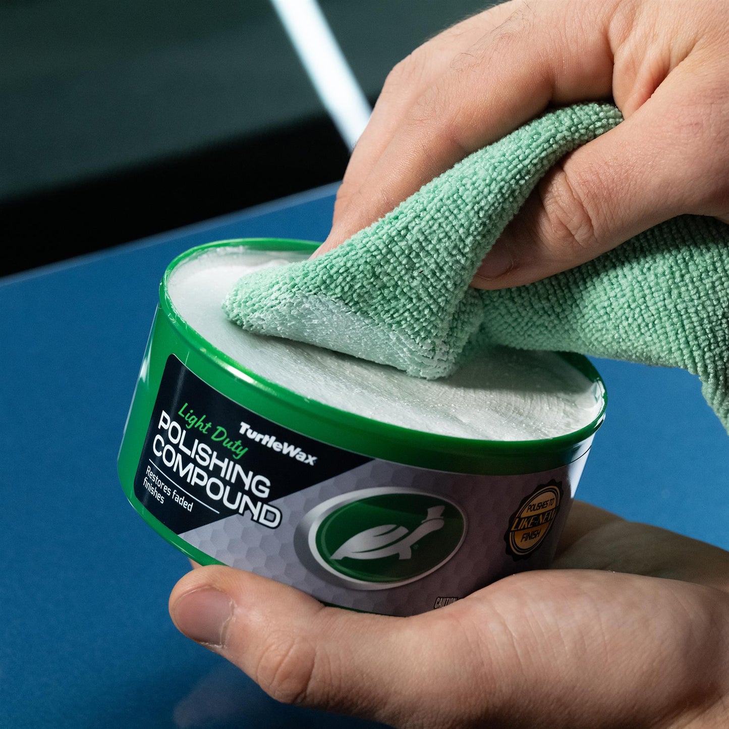 Hands applying Turtle Wax polishing compound with a microfiber cloth on a car surface. The compound restores faded finishes.